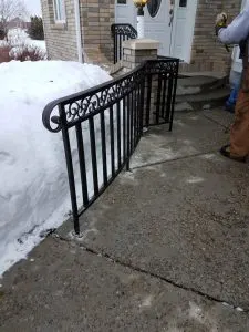 Black ornamental metal handrail with scrollwork on snowy concrete front steps