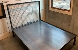 Welded steel platform bed frame with wire mesh headboard against a concrete wall